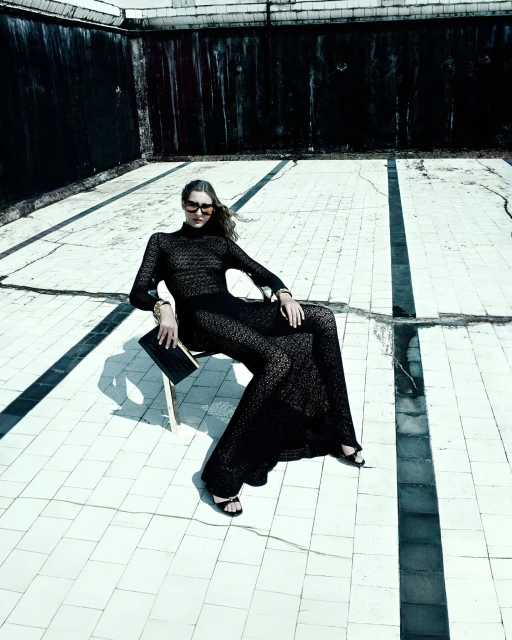 A woman in a black lace dress sitting on a chair in an empty swimming pool wearing sunglasses and holding a black clutch.