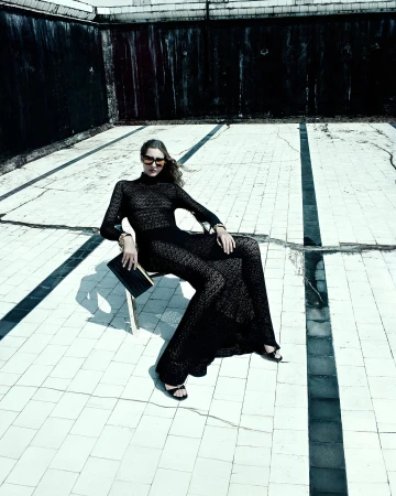 A woman in a black lace dress sitting on a chair in an empty swimming pool wearing sunglasses and holding a black clutch.