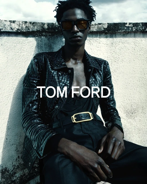 Fashion shot of a dark-skinned male model wearing sunglasses, black jacket, black shirt, and black pants against a white concrete wall.
