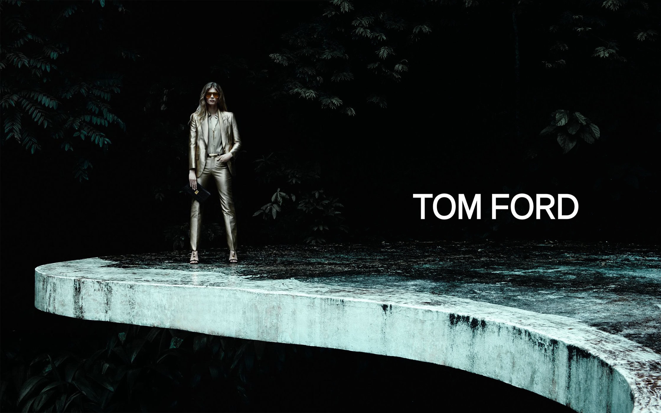 A woman in a gold suit stands on a curved concrete surface in a fashion shot with trees in the background and 'TOM FORD' text.