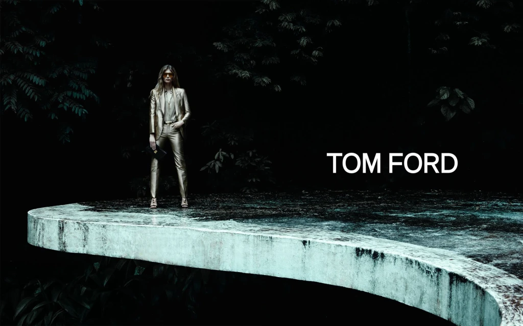 A woman in a gold suit stands on a curved concrete surface in a fashion shot with trees in the background and 'TOM FORD' text.