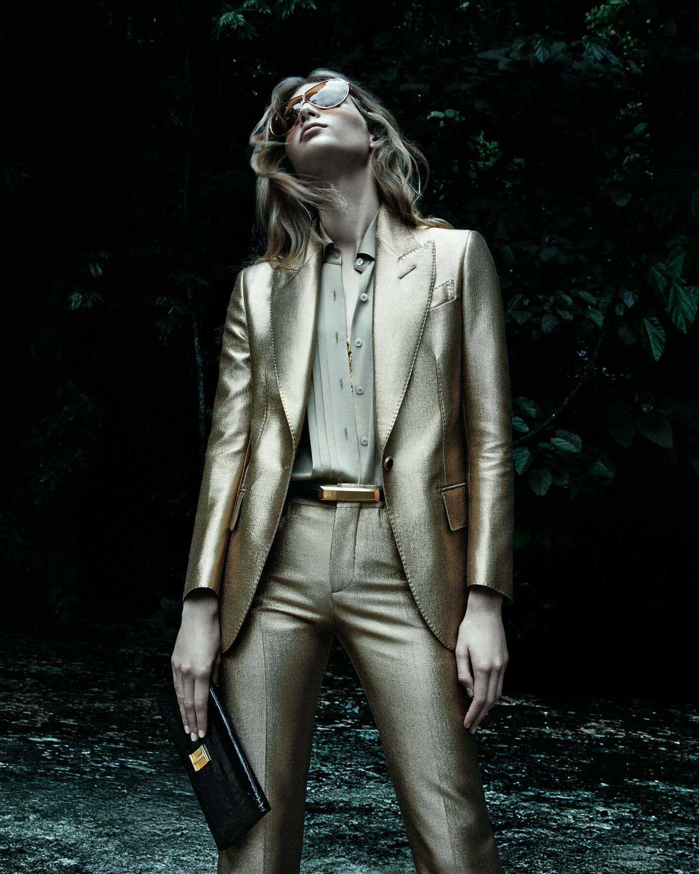 Model poses in a gold metallic suit and sunglasses in a dark, outdoor setting, holding a black clutch, looking upward in a fashion shot.