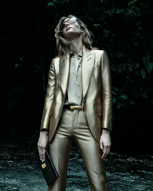 Model poses in a gold metallic suit and sunglasses in a dark, outdoor setting, holding a black clutch, looking upward in a fashion shot.