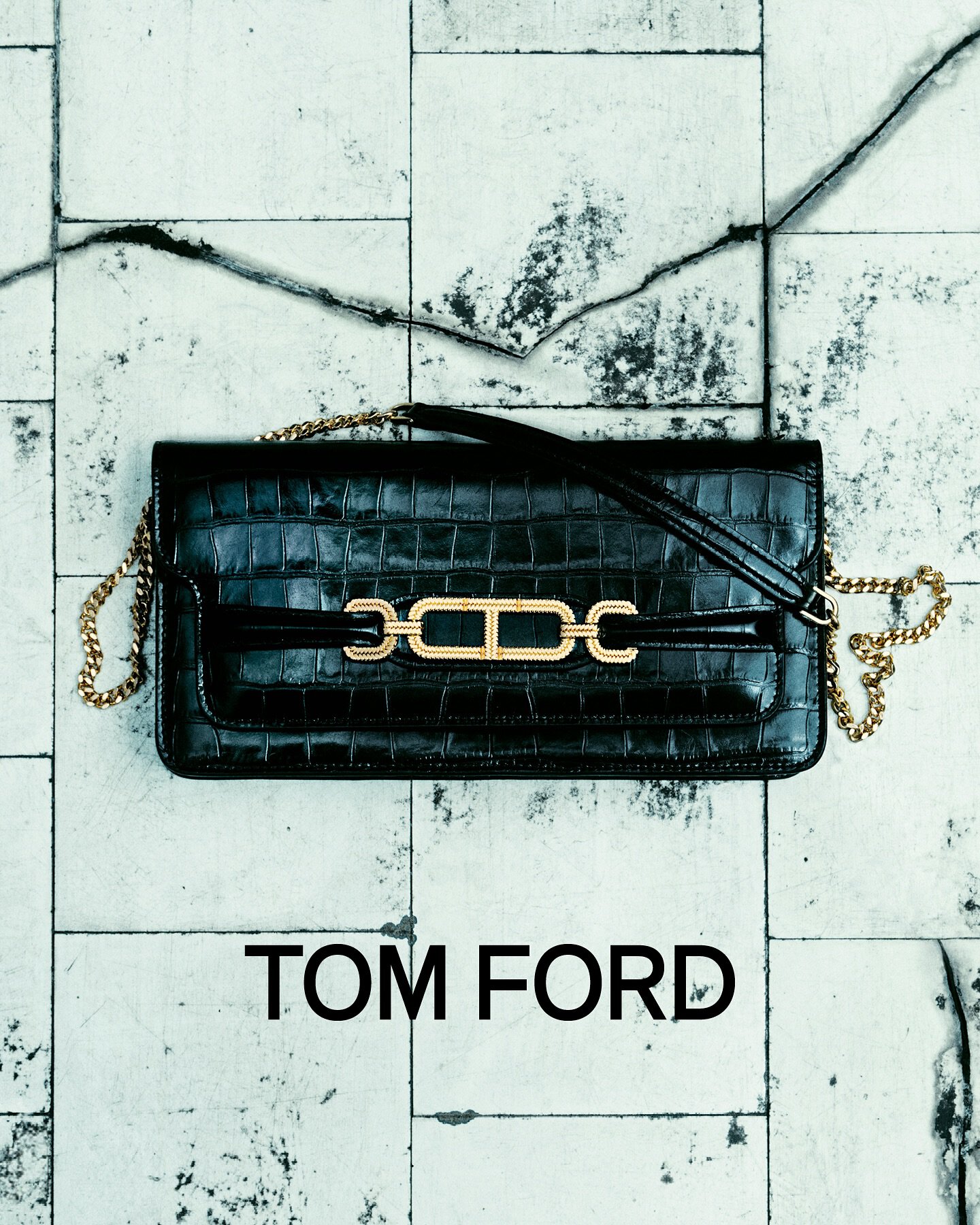 Stylish black leather Tom Ford handbag with gold chain strap and hardware accent on a textured white tile surface. Fashion product shot.