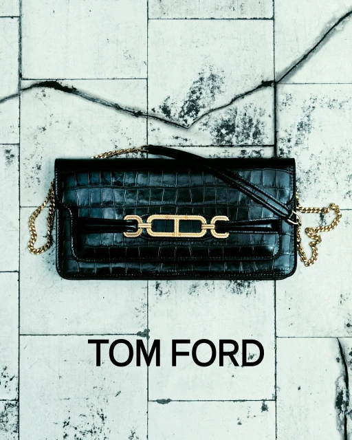 Stylish black leather Tom Ford handbag with gold chain strap and hardware accent on a textured white tile surface. Fashion product shot.
