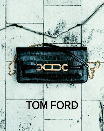 Stylish black leather Tom Ford handbag with gold chain strap and hardware accent on a textured white tile surface. Fashion product shot.