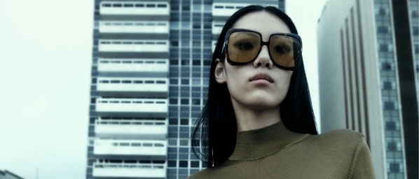 Model wearing oversized tortoiseshell sunglasses and a fitted olive top against a tall urban building exterior