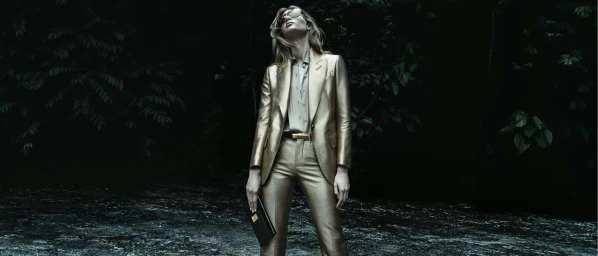 Model in a metallic gold suit and sunglasses holding a black clutch standing before a dark forest