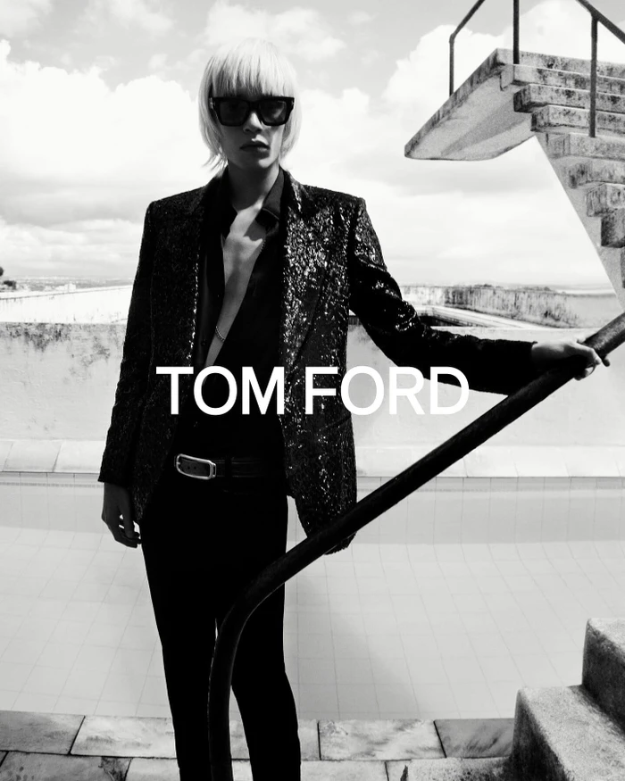 Model in sunglasses and sequin blazer standing by a rooftop pool under an overcast sky.
