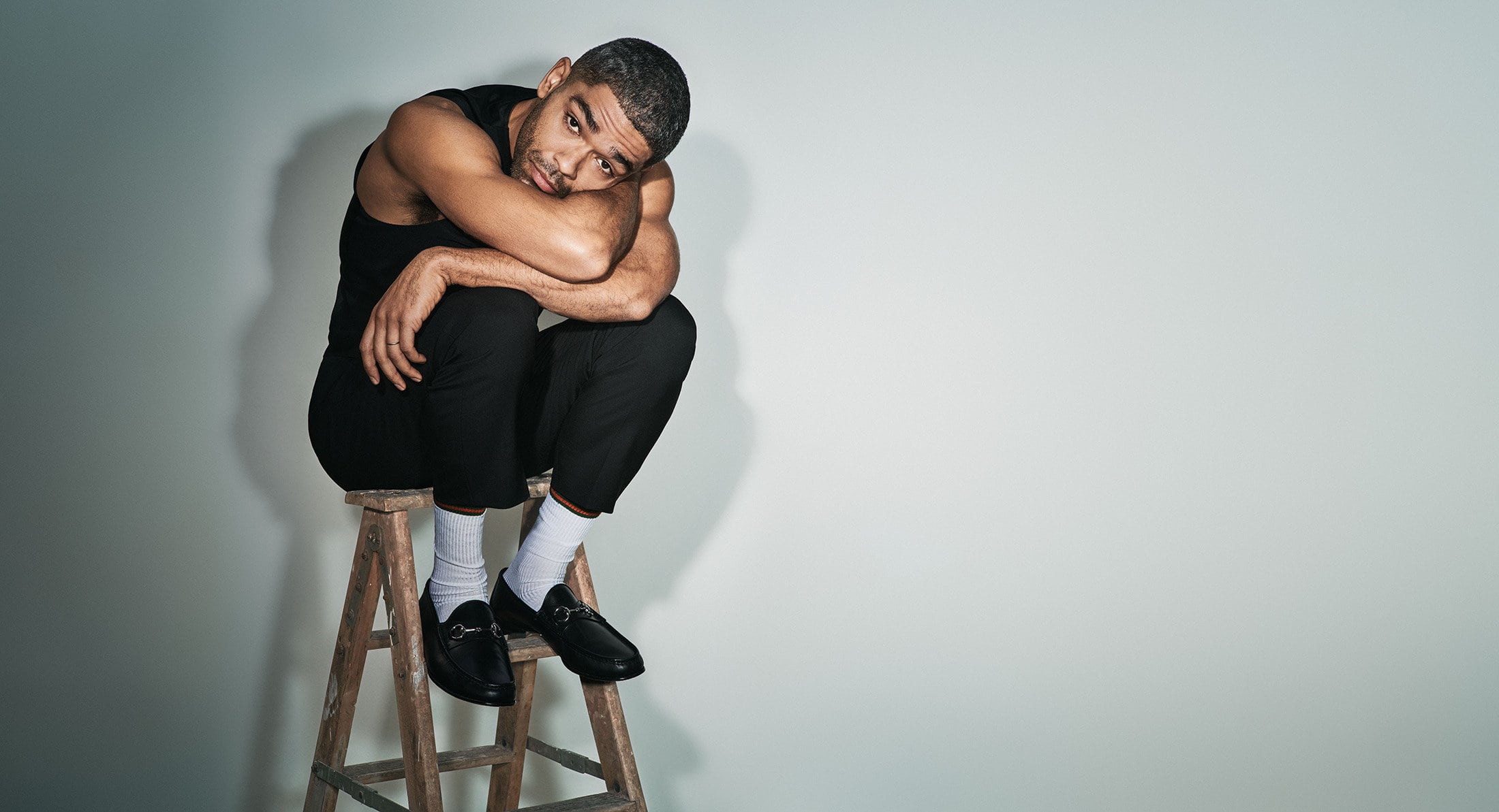 Portrait of a man wearing a black tank top and pants sitting on a wooden step stool in a studio with a neutral grey background.