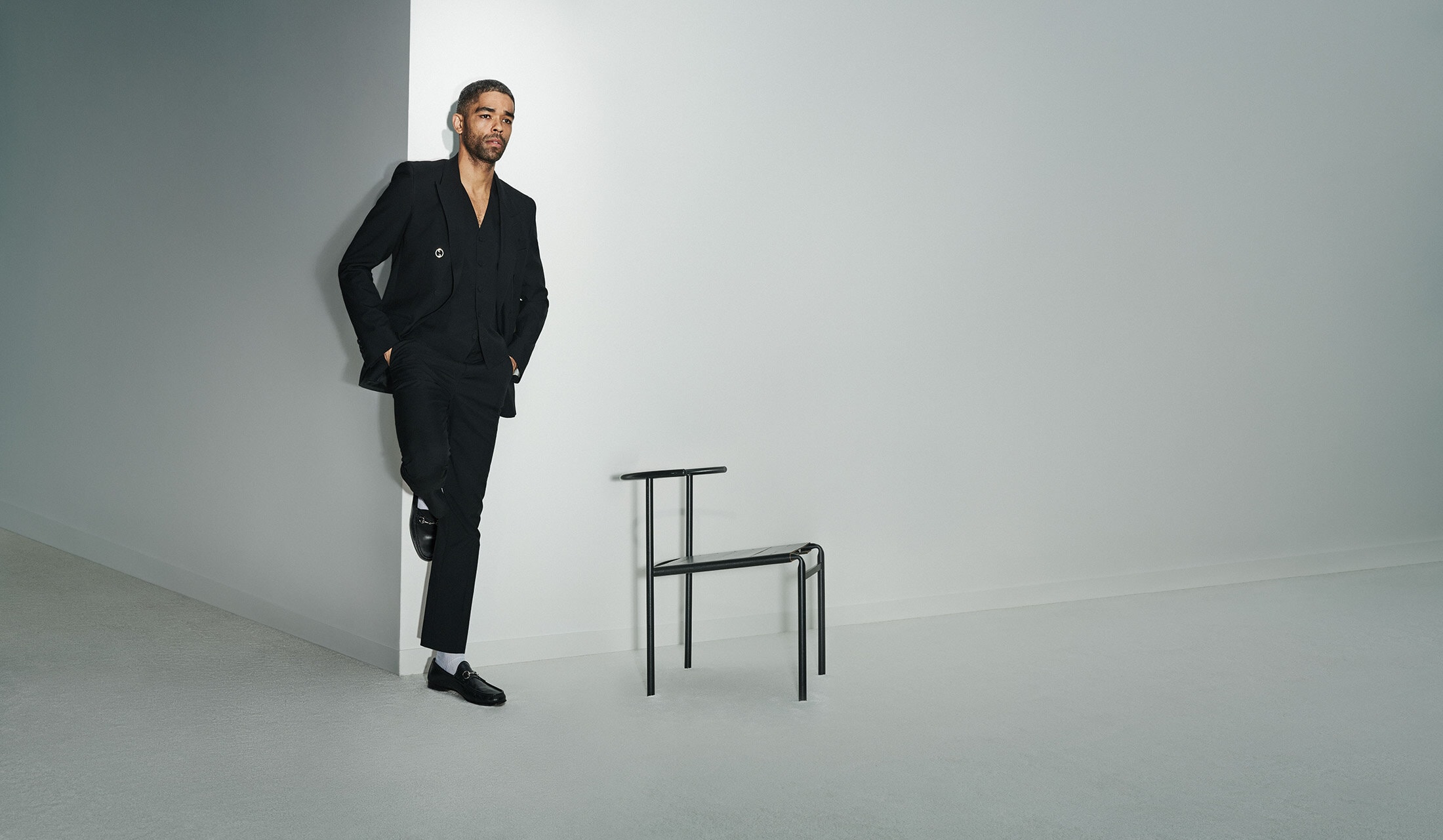 A Black man in a stylish black suit leans against a white wall next to a minimalist chair in a brightly lit indoor setting.