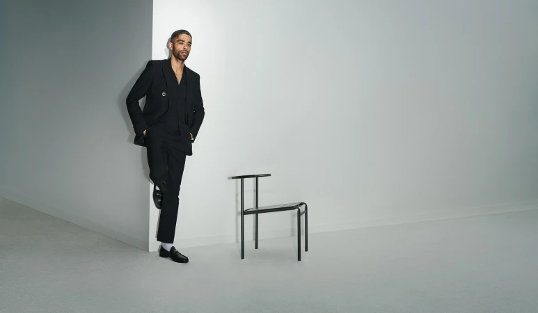 A Black man in a stylish black suit leans against a white wall next to a minimalist chair in a brightly lit indoor setting.