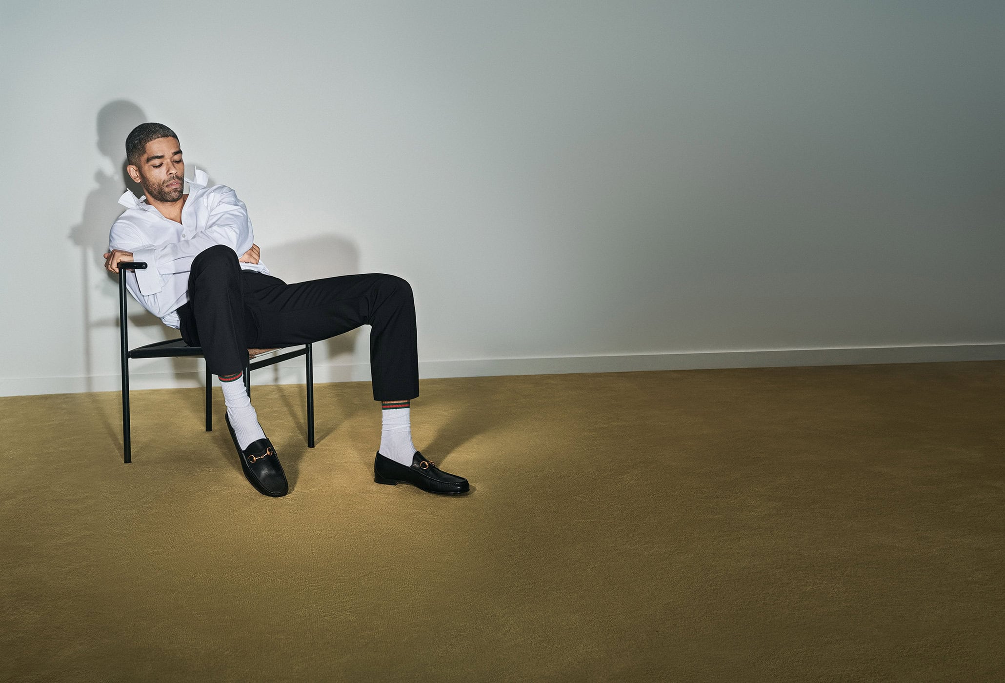 Adult male seated in a minimalist indoor setting, wearing a white shirt, dark pants, white socks, and loafers, with a neutral expression.