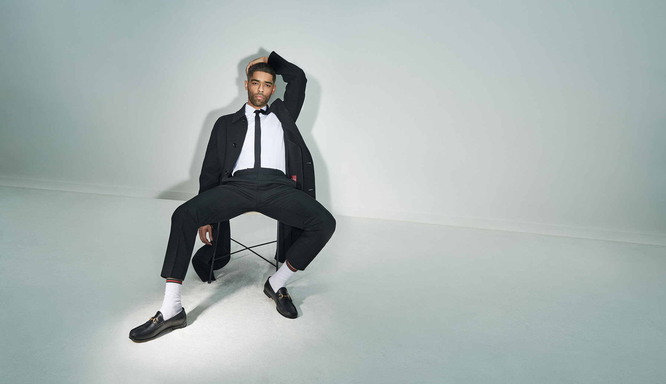 An adult male sits on a chair in a photo studio wearing a black suit and white socks, holding a hat, and posing with his hand on his head.