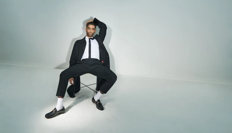 An adult male sits on a chair in a photo studio wearing a black suit and white socks, holding a hat, and posing with his hand on his head.