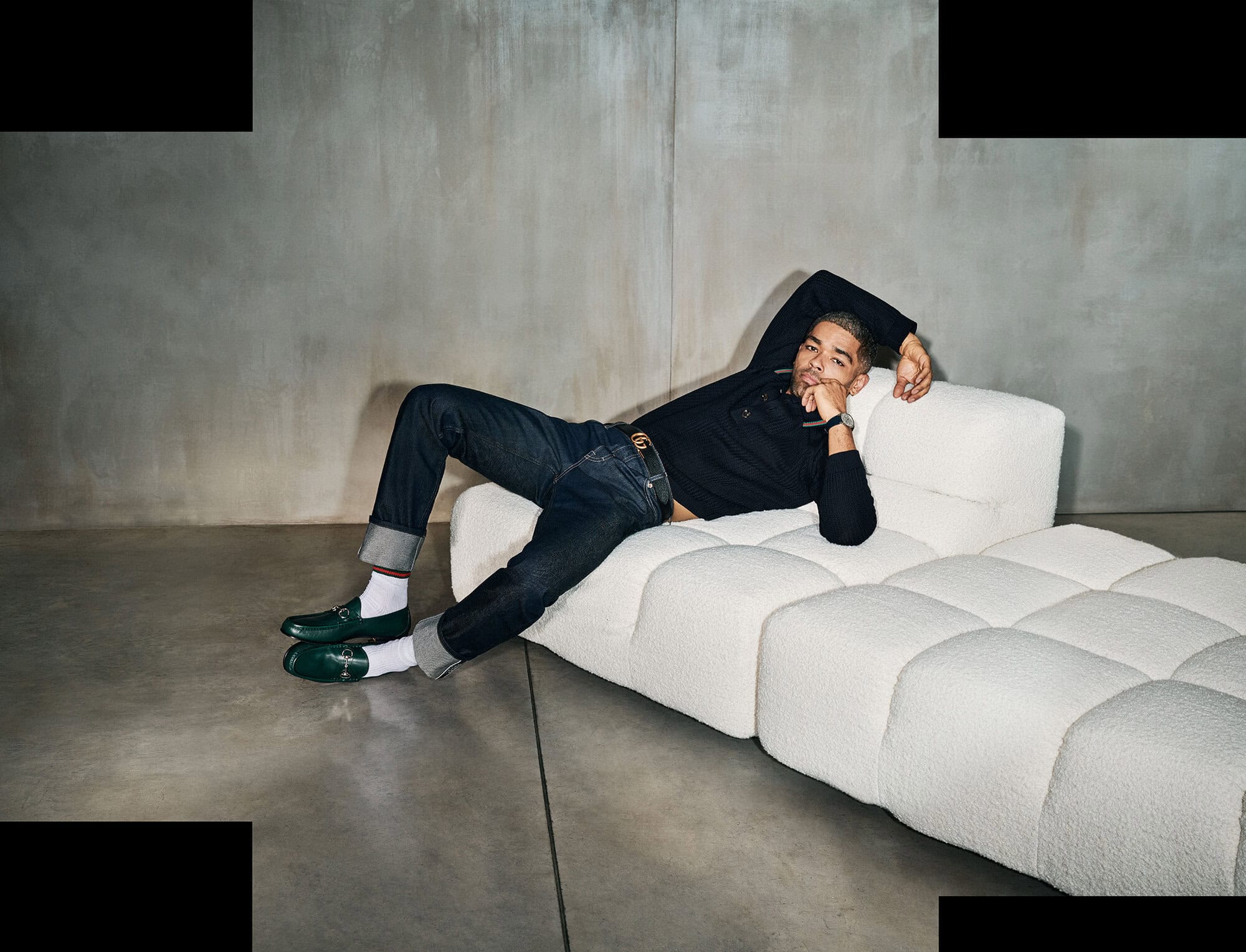 Man in dark denim and a black sweater reclines on a white modern sofa against a neutral wall in a studio setting with green loafers.