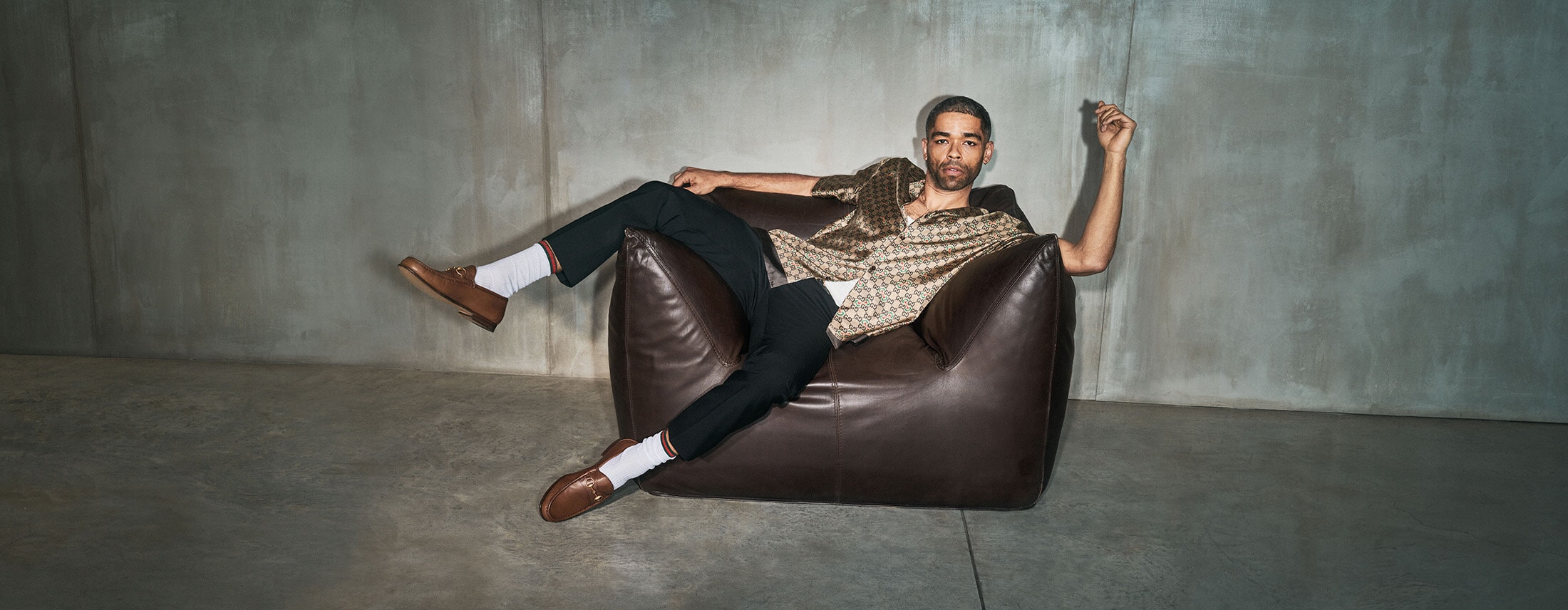 Adult male poses in a patterned shirt and dress pants on a leather beanbag chair in an indoor studio with a grey concrete background.