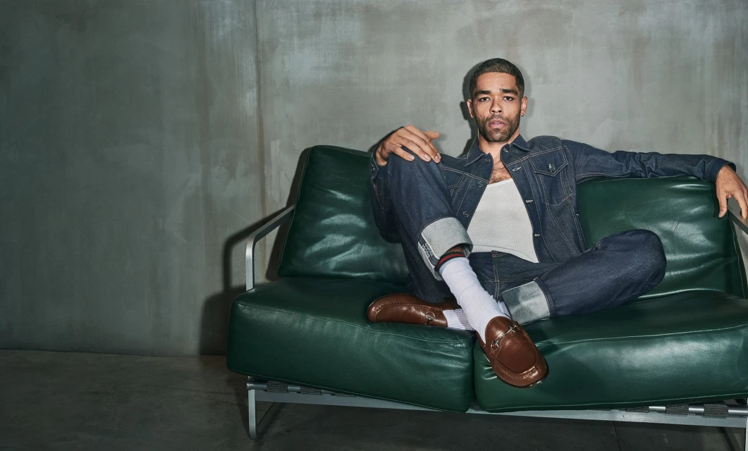 A man in a denim jacket and jeans sits casually on a green leather couch in a studio, illuminated by natural light against a grey backdrop.