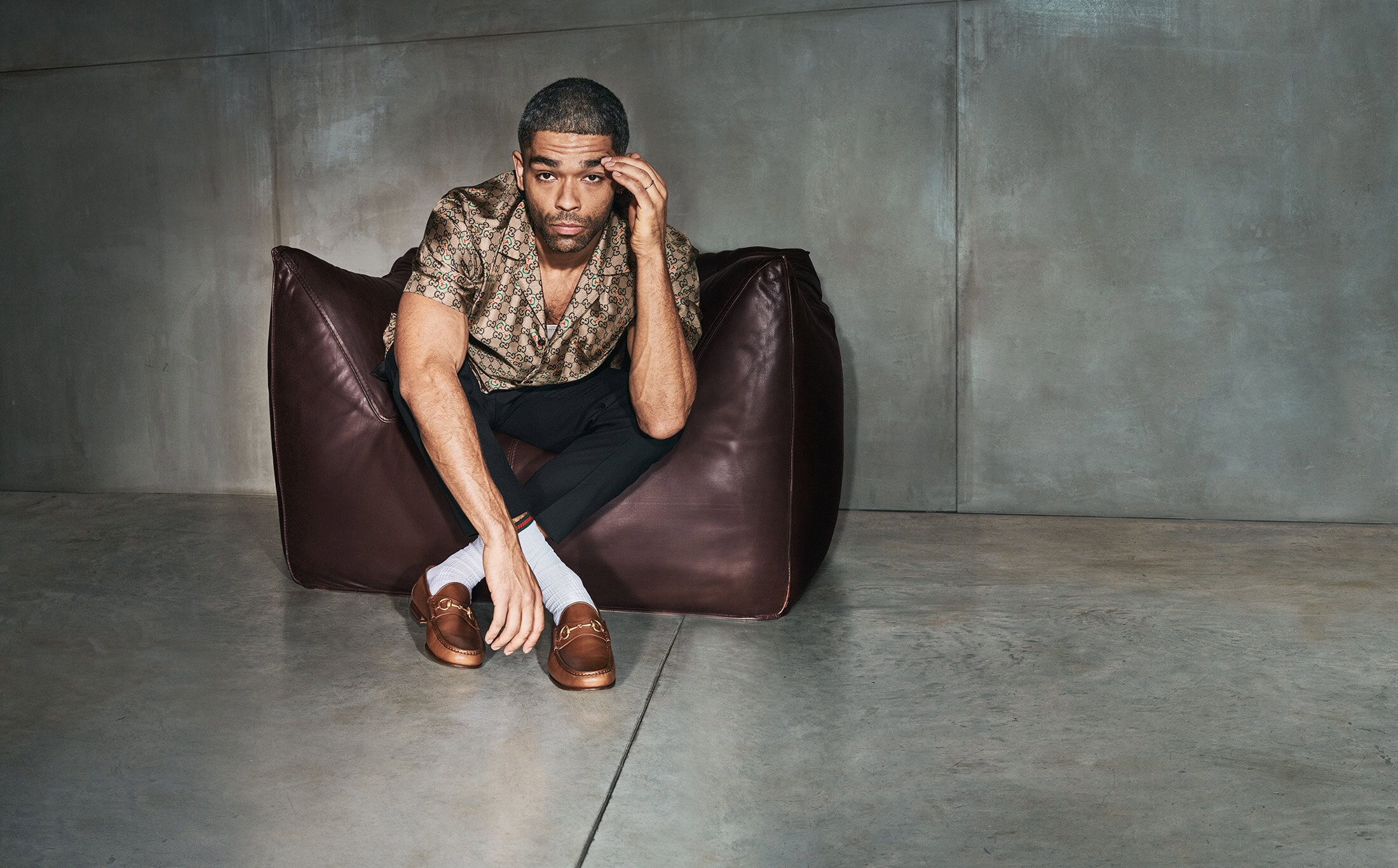 A man with short hair sitting in a brown leather chair, wearing a patterned shirt, black pants, white socks, and brown loafers indoors.
