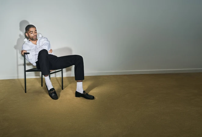 Kingsley Ben-Adir seated in a black chair wearing a white shirt and black loafers for Gucci