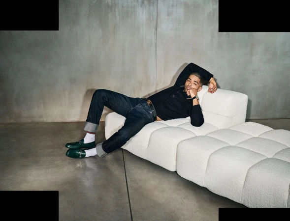 Kingsley Ben-Adir reclining on a white tufted sofa wearing green loafers and dark jeans for Gucci.