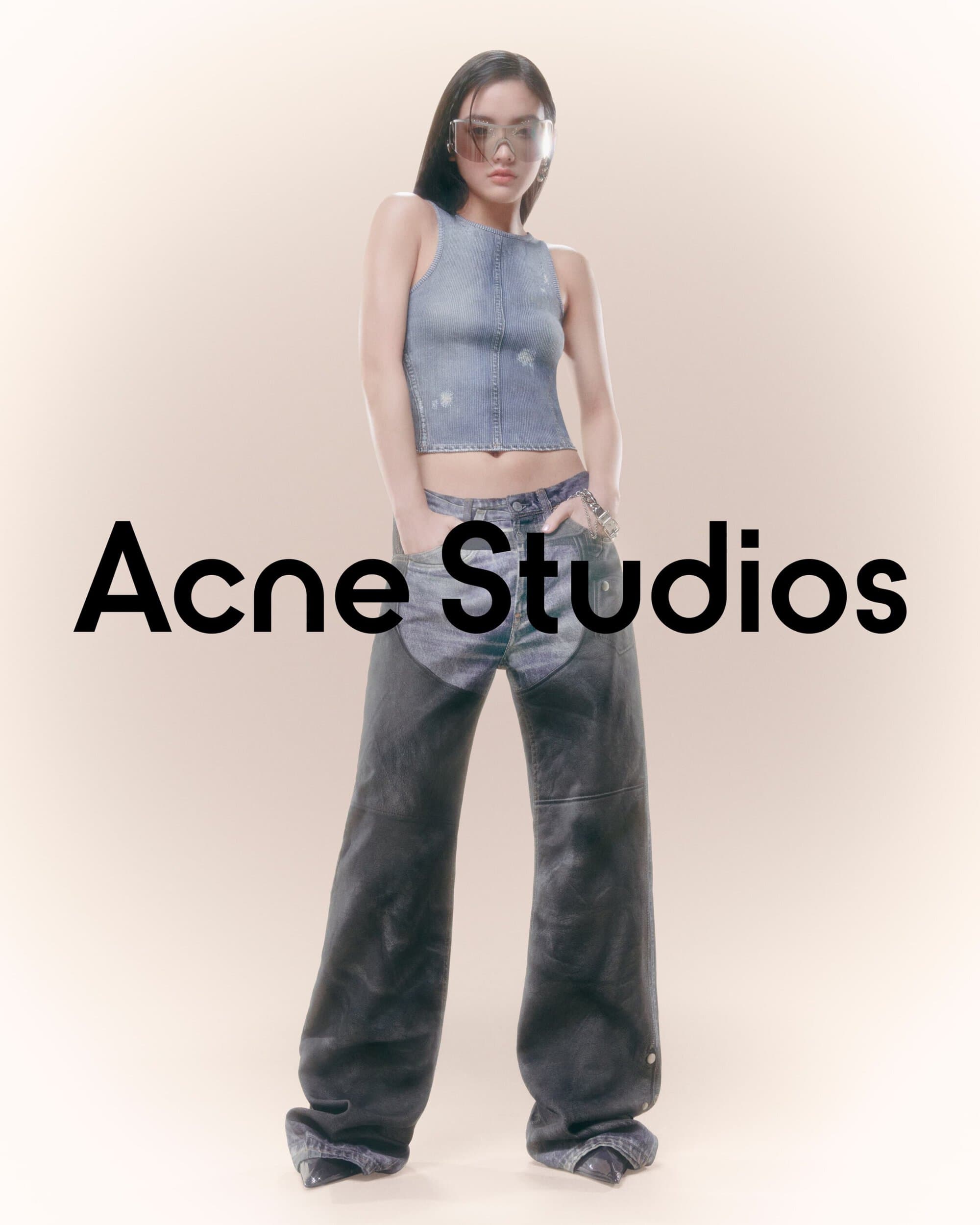 Fashion model wears denim top and baggy jeans by Acne Studios in a studio shot. The model accessorizes with sunglasses and a bracelet.