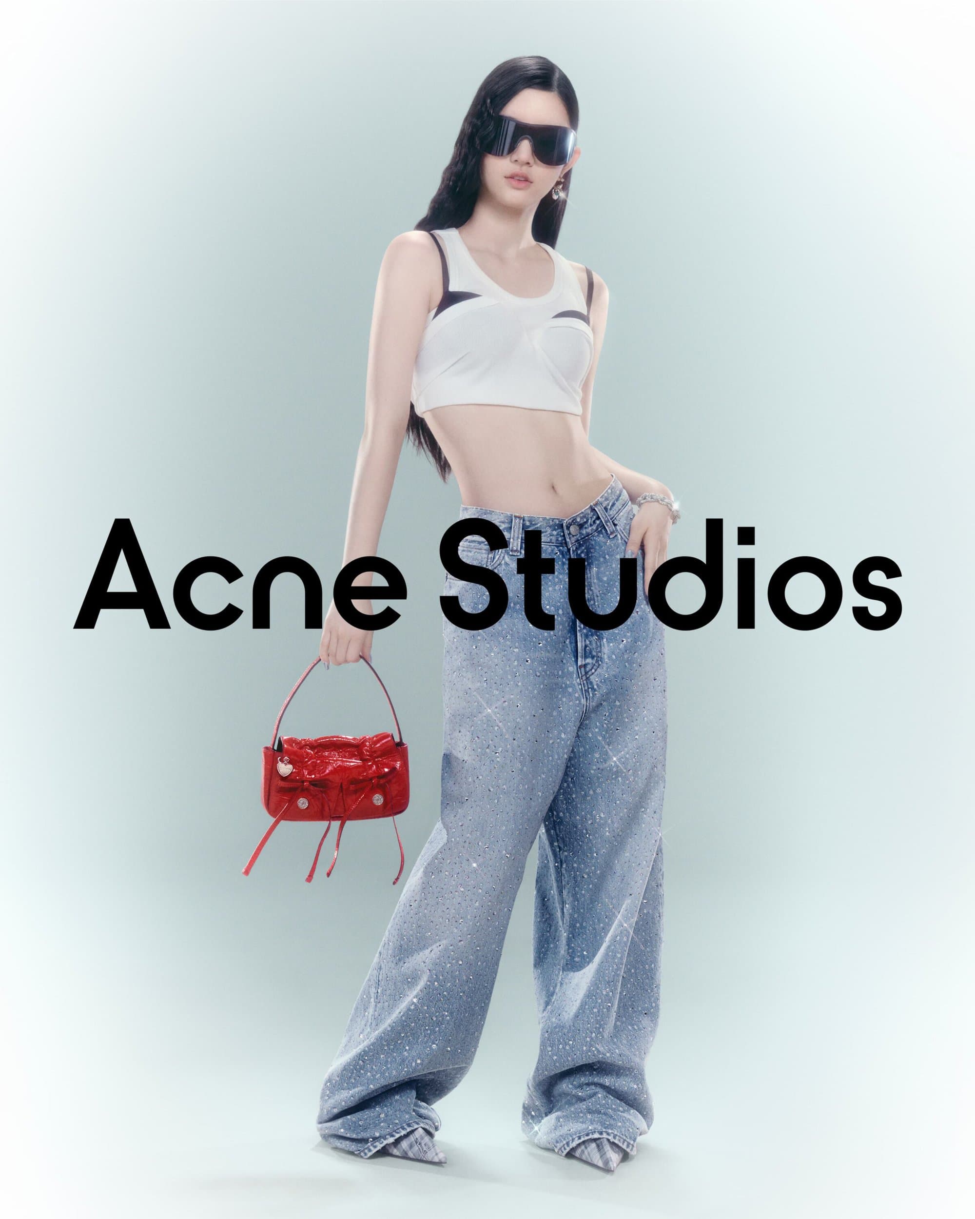 A fashionable woman poses in a light studio wearing a white crop top, wide leg jeans and sunglasses, holding a red handbag.
