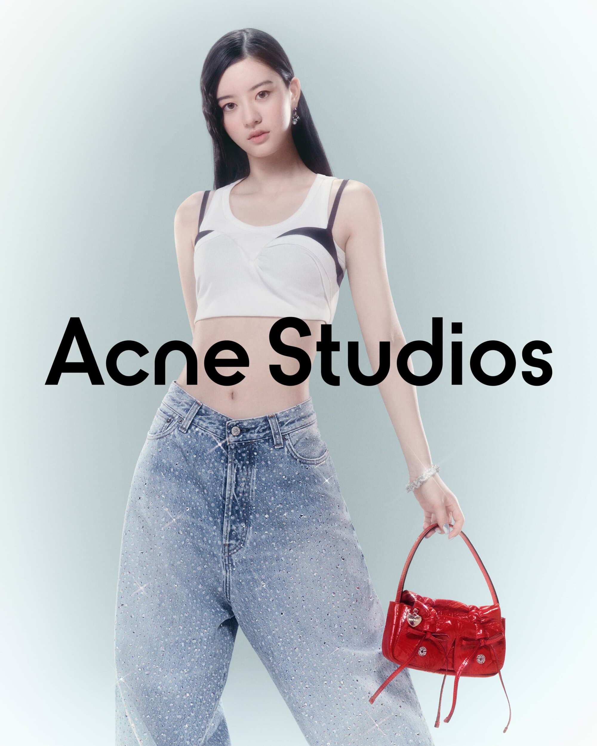 A young woman in a white crop top and embellished jeans poses indoors with a red purse in a fashion-forward editorial shot.