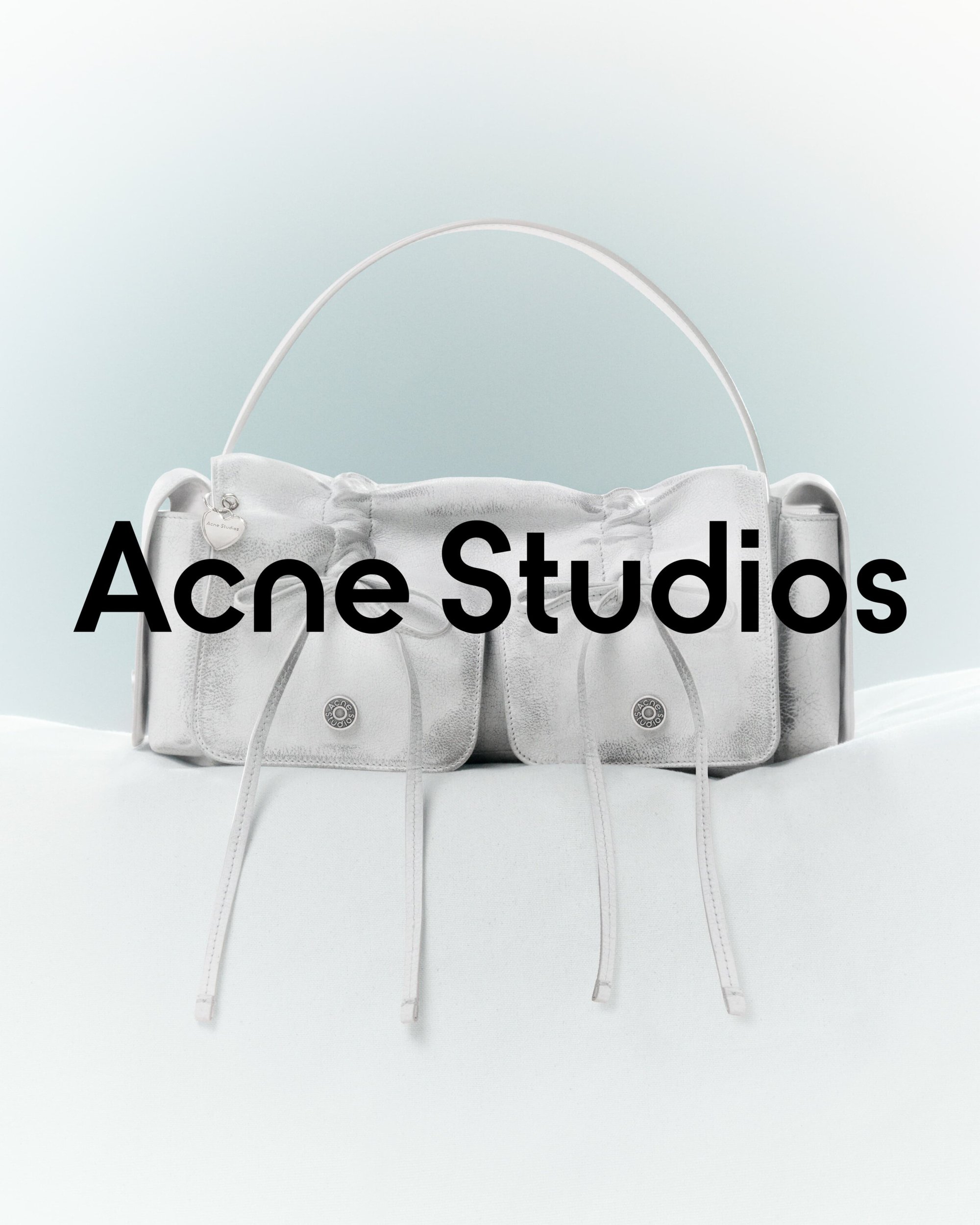 Silver-toned Acne Studios handbag on a light surface against a pale gradient background, emphasizing its design and texture.