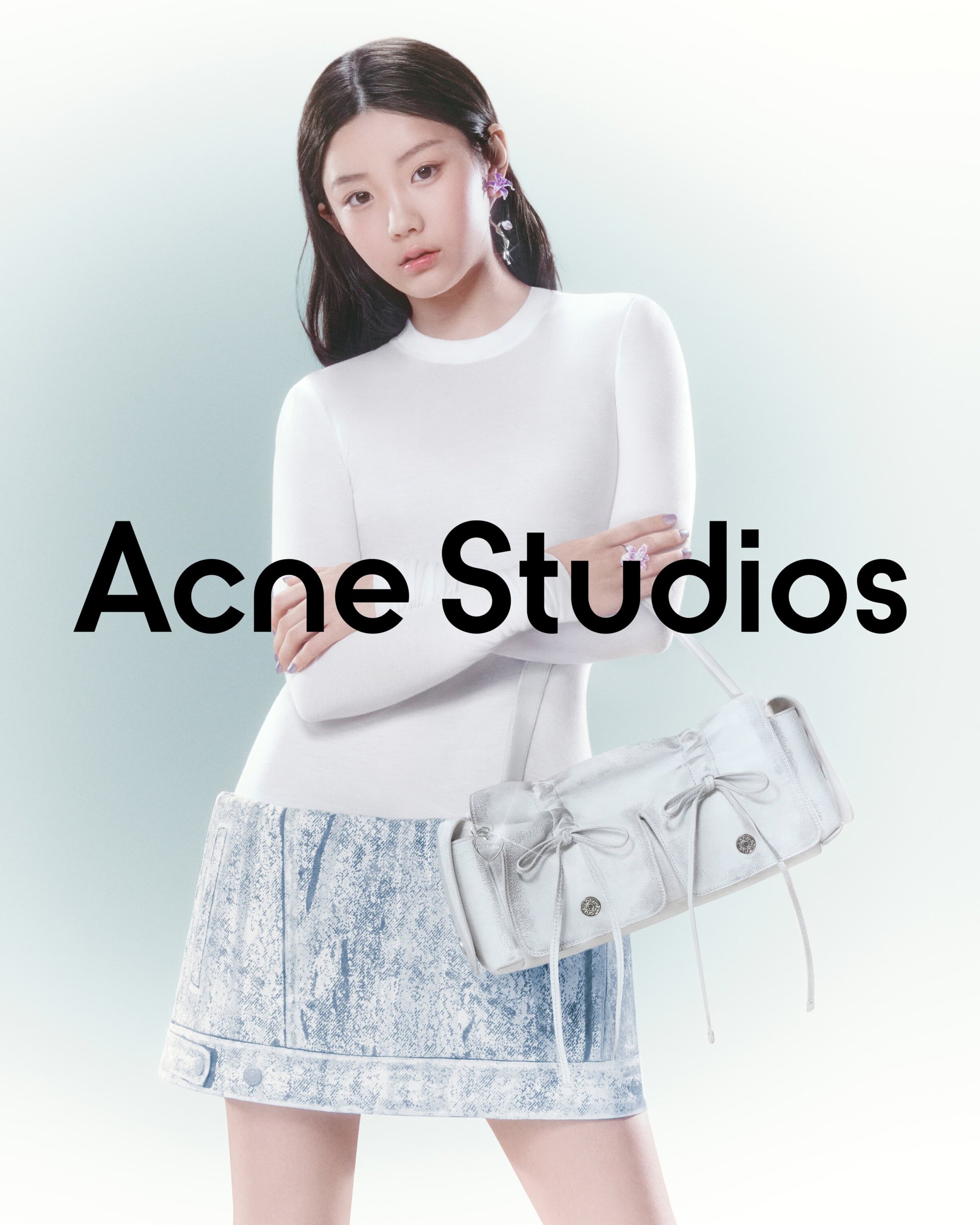 An Asian female model poses with a light-blue denim skirt and a white leather handbag with white drawstrings for Acne Studios.