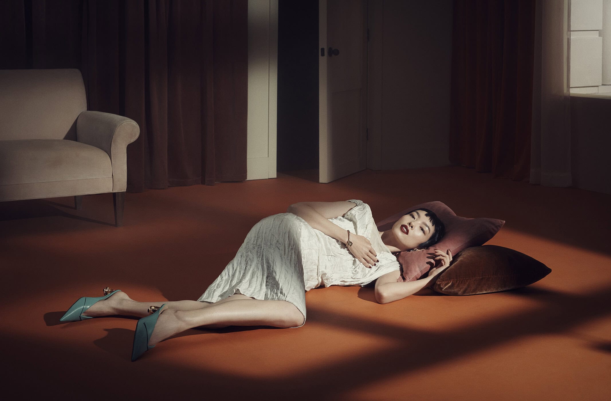 Adult female with dark hair and dark lipstick in white dress and blue heels laying on a pillow in a staged indoor setting.