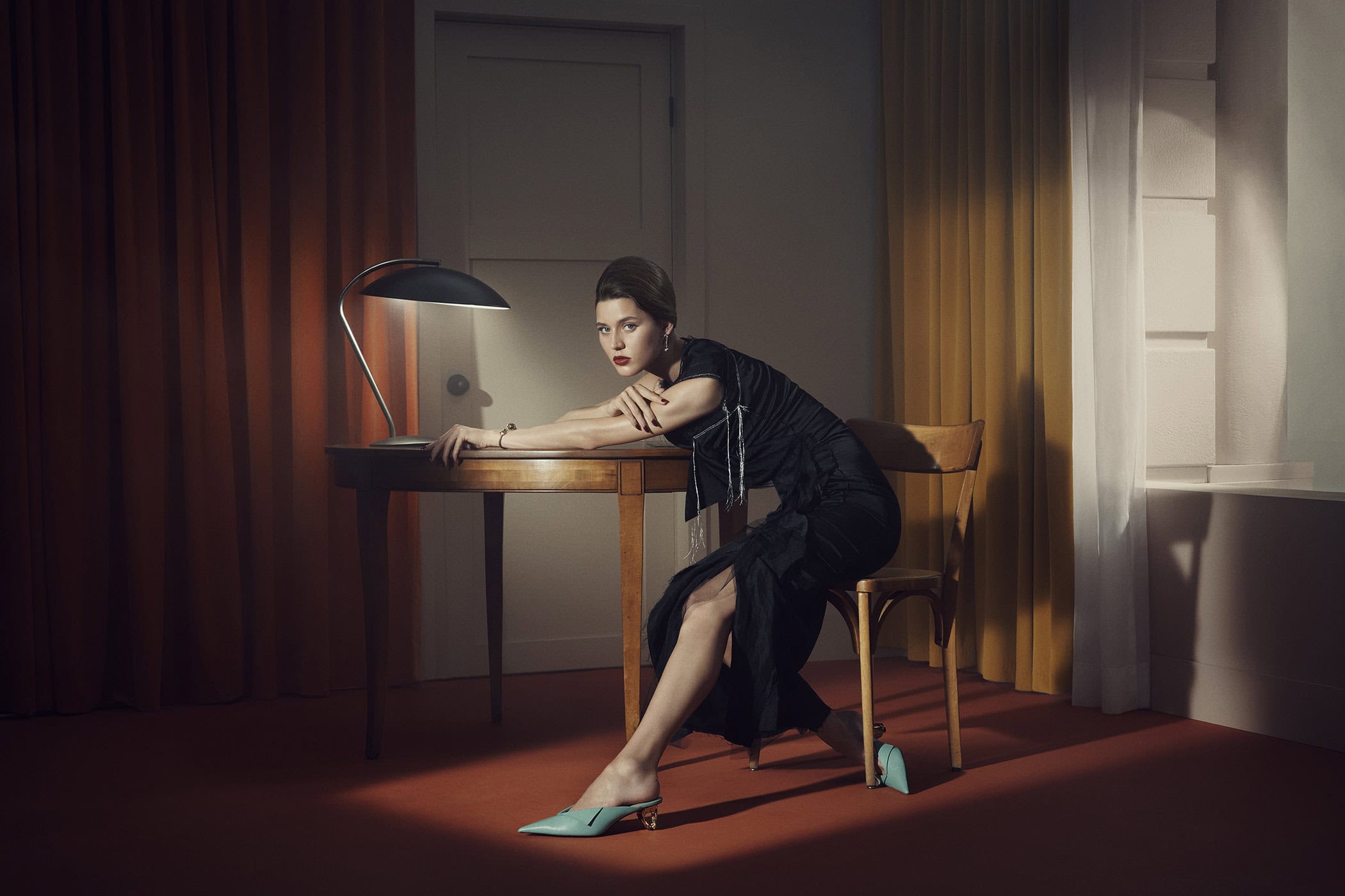 Elegant woman in black dress sits at desk in a warm-toned room with curtains and vintage-style lamp, showcasing fashion and interior.