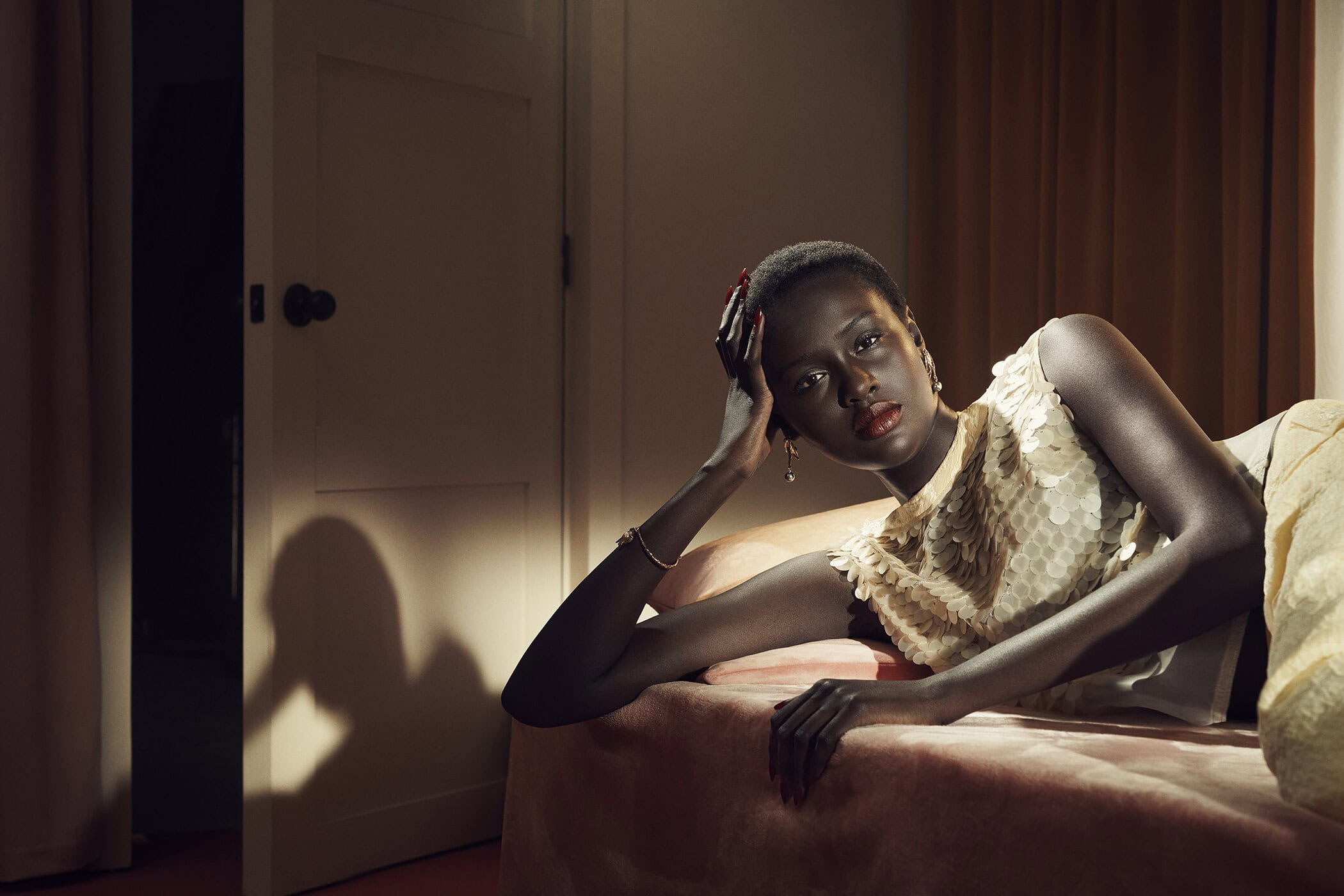 Elegant woman lies on a pink bed, wearing a sequined top in a softly lit room with a white door and warm brown curtains, portrait orientation.