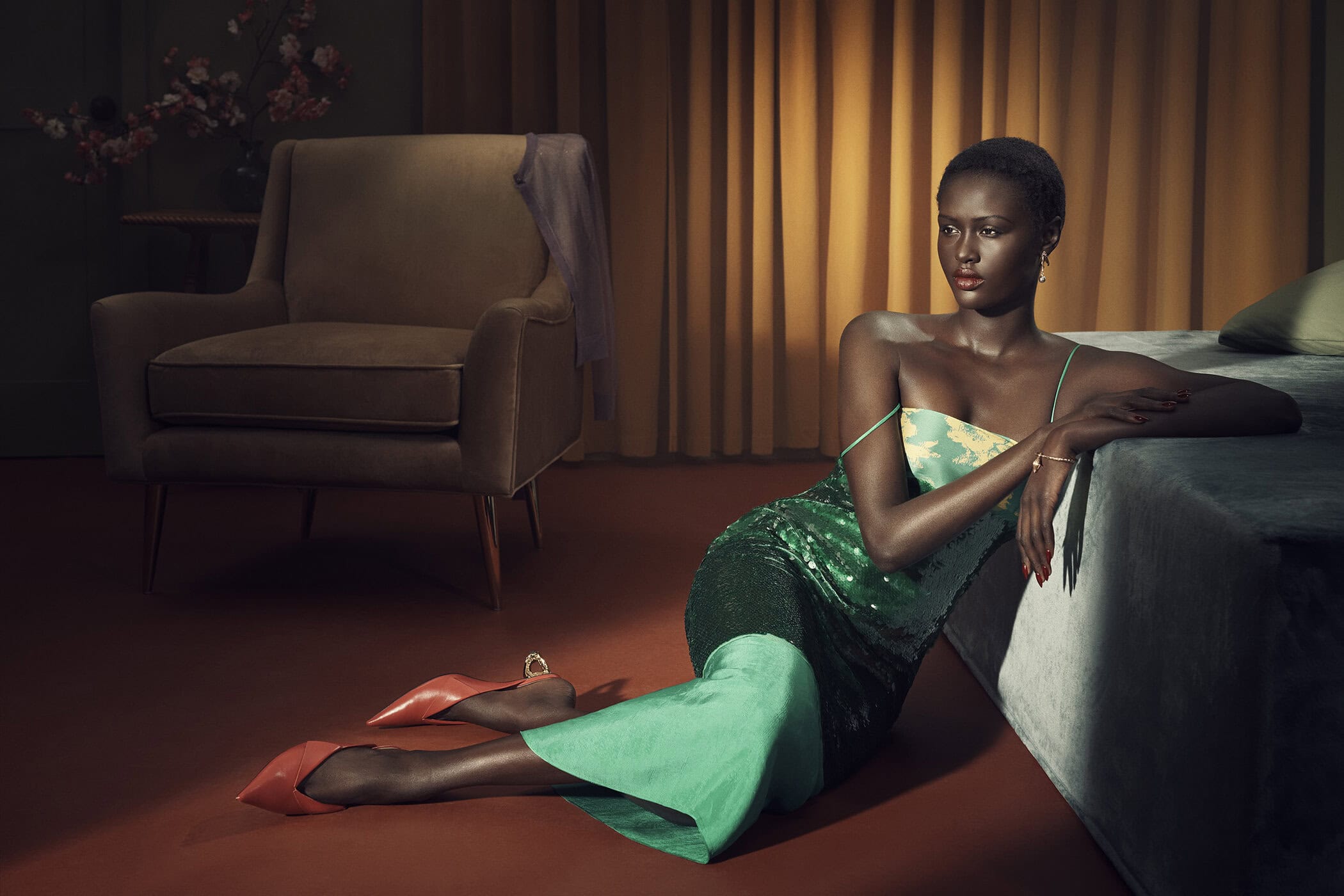 A seated black woman models a green sequin dress in a vintage-inspired interior setting with an armchair, curtains, and a bed.