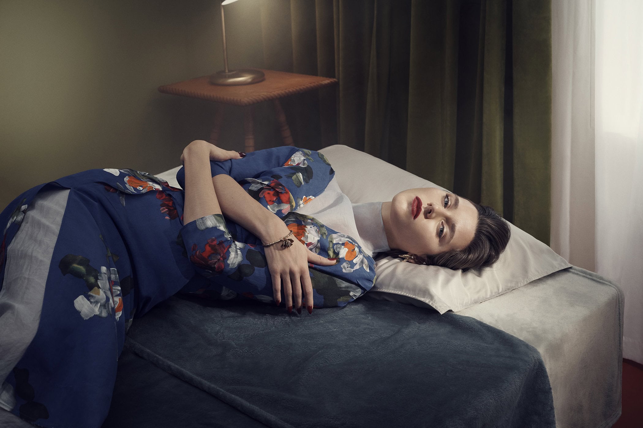 Adult woman lies on a bed in a stylish, floral blue dress with a white collar, accessorized with bracelet, in an indoor setting.