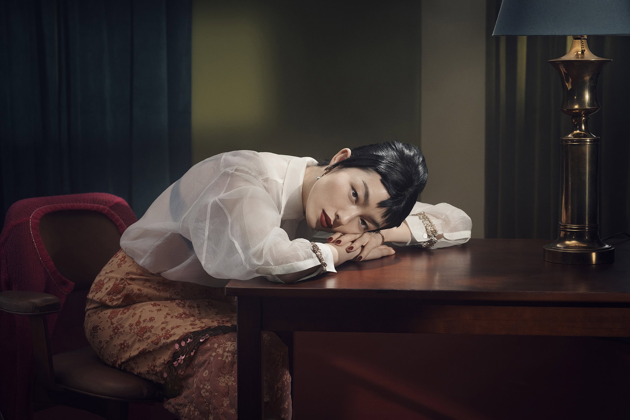 A woman rests her head on a wooden desk, wearing a sheer white blouse and floral skirt, with a lamp and curtains in the background.