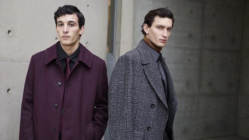 Two adult men wearing stylish overcoats pose outdoors against a concrete wall in a medium shot, showcasing fashion and style.