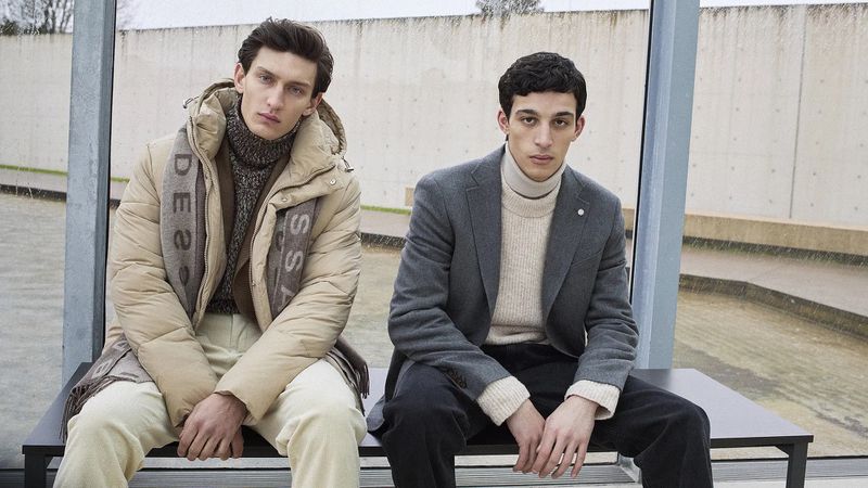 Two adult males model stylish fall fashion. One wears a puffer jacket and scarf, the other a blazer, seated on a bench outdoors.