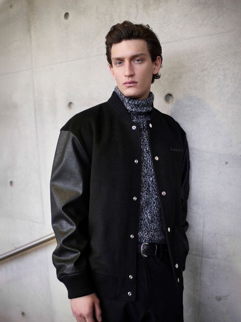 A stylish young man in a black letterman jacket and gray turtleneck poses against a concrete wall, showcasing a blend of casual and chic.