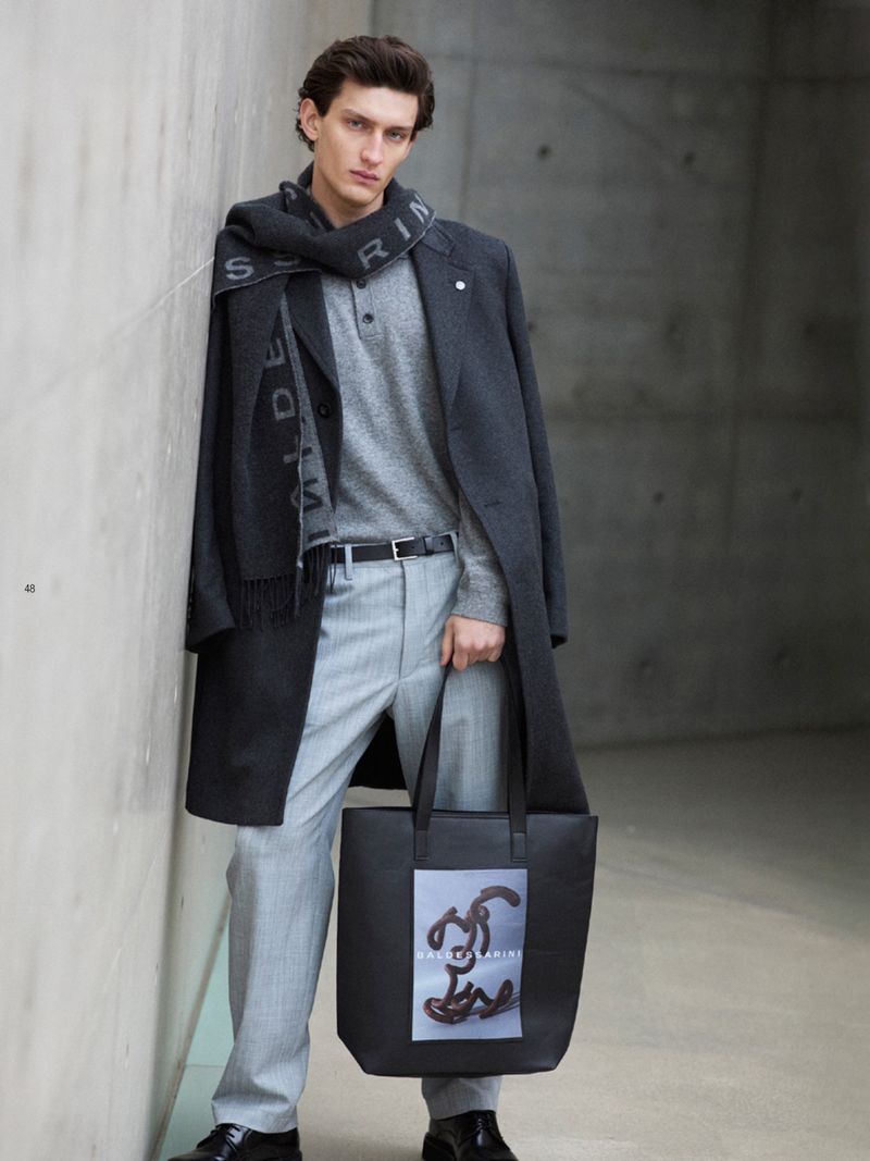Male model leans against a grey wall wearing a grey coat, light grey trousers, and a scarf, holding a black tote bag.