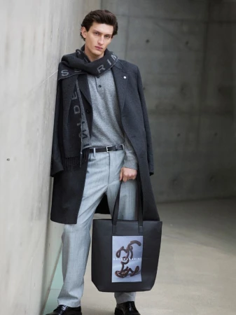 Male model leans against a grey wall wearing a grey coat, light grey trousers, and a scarf, holding a black tote bag.