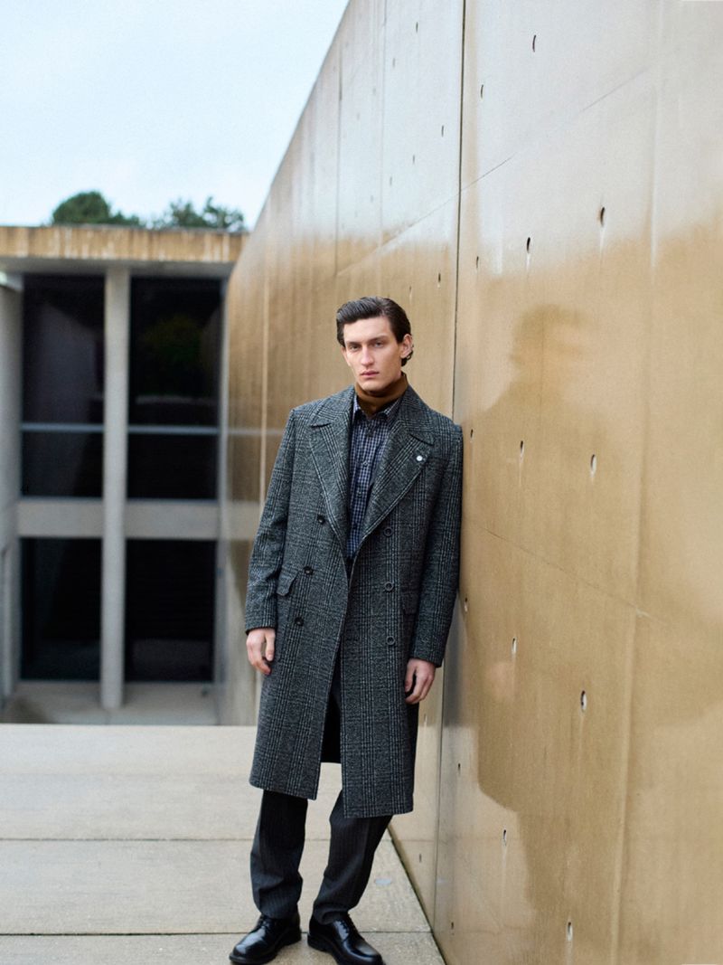 Man in gray overcoat leaning against a tan concrete wall of a building with large windows, creating a stylish outdoor portrait.