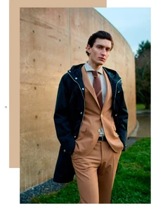 Man wearing a camel suit, brown tie, and black raincoat standing outdoors near a curved concrete wall and lawn.