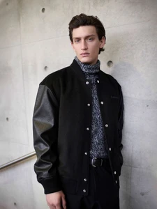 Model wearing a black varsity jacket and grey turtleneck leaning against a concrete wall for Baldessarini