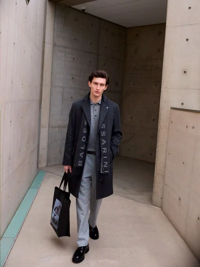 Model in a dark wool coat and grey trousers walking outdoors in a concrete architectural corridor