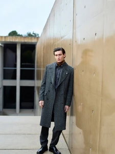 Man wearing a long grey tweed coat leaning against a smooth concrete wall outdoors