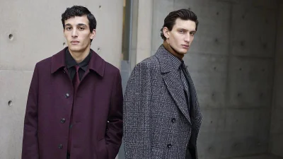 Two men standing against a textured grey concrete wall, wearing tailored coats and shirts for the Baldessarini F/W 2025 campaign.