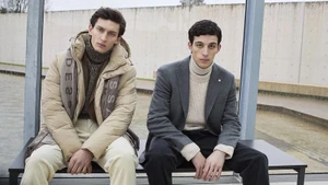 Two men seated on a bench wearing winter outerwear including a beige puffer jacket and grey blazer for Baldessarini.