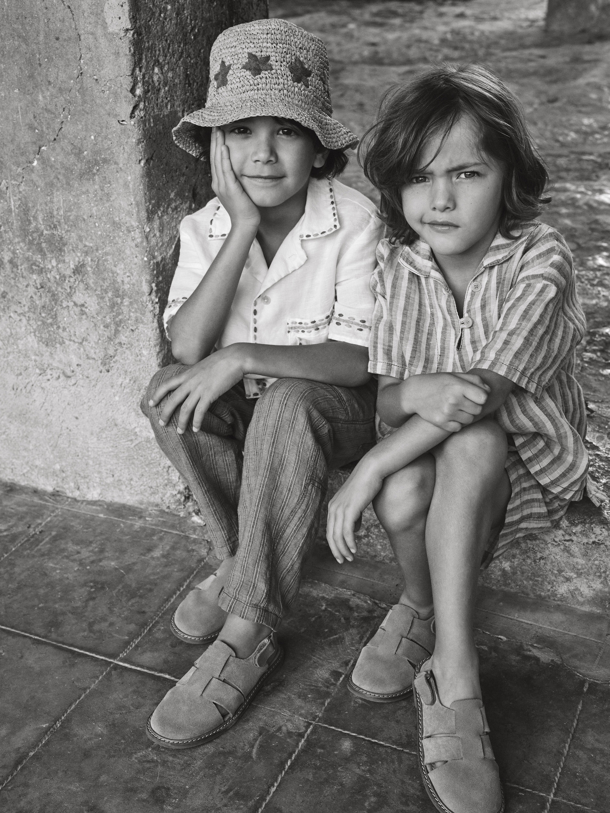 Two children sit together in a vintage-style black and white image. One wears a straw hat, while both wear casual clothing and sandals.