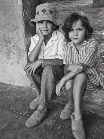 Two children sit together in a vintage-style black and white image. One wears a straw hat, while both wear casual clothing and sandals.